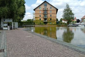 En byggnad av tegel med en balkong, en stenlagd gångväg och en vattenyta i förgrunden.