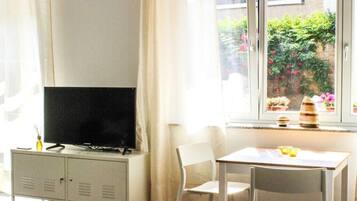 Ein Zimmer mit Fernseher auf einem Schrank, einem Tisch mit Stühlen und einem Fenster mit Blick auf Grünflächen.