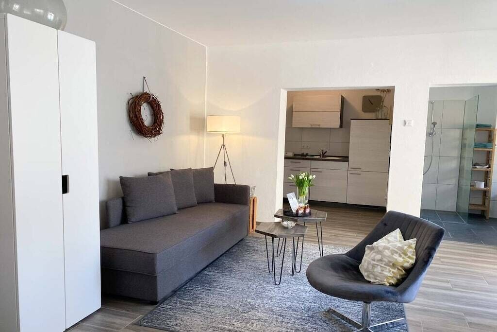 Ein modernes Wohnzimmer mit einem grauen Sofa, einem kleinen runden Tisch und einem dunklen Sessel. Im Hintergrund ist eine Küche zu sehen.