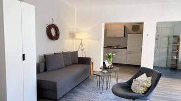 Ein modernes Wohnzimmer mit einem grauen Sofa, einem kleinen runden Tisch und einem dunklen Sessel. Im Hintergrund ist eine Küche zu sehen.