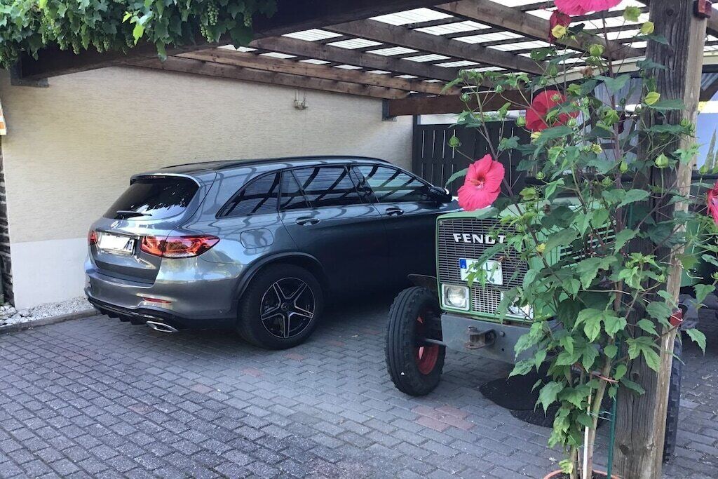 Ein geparktes SUV in einem überdachten Parkbereich, mit einem Gartenschlauch und einer Pflanze mit roten Blüten.