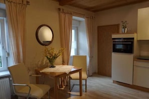 Cuina compacta amb zona de dinar, taula de fusta, cadires, nevera petita i finestra amb cortines.