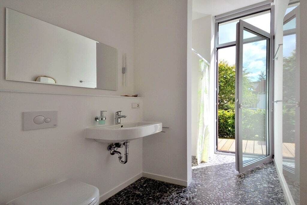Casa de banho com lavatório branco, sanita, espelho e porta de vidro que dá para zona exterior.