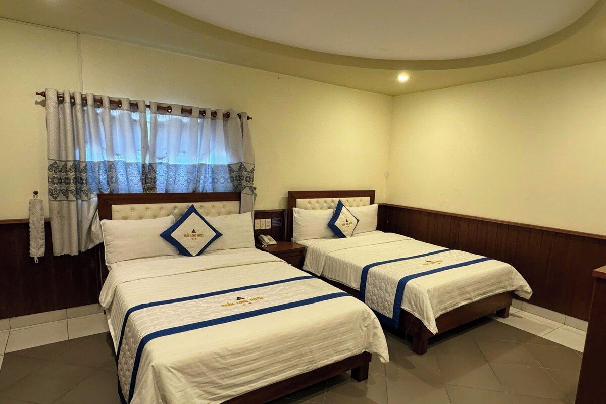 Una habitación de hotel con dos camas, una mesita de noche, un teléfono y una ventana con cortinas.