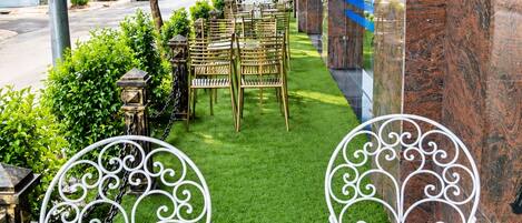 An outdoor seating area with white metal chairs and tables, surrounded by greenery and a view of the street.