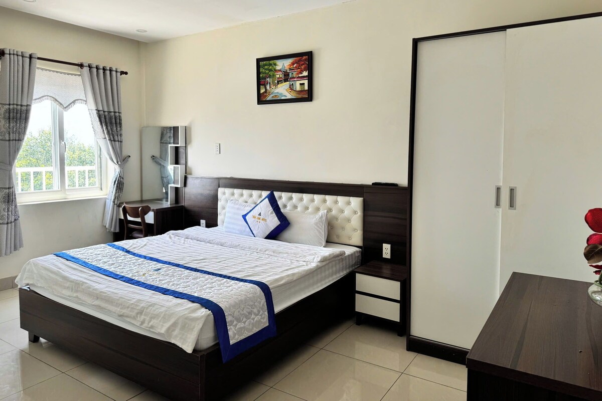 Habitación de hotel con cama, mesitas de noche, armario, un cuadro en la pared y ventana con cortinas.