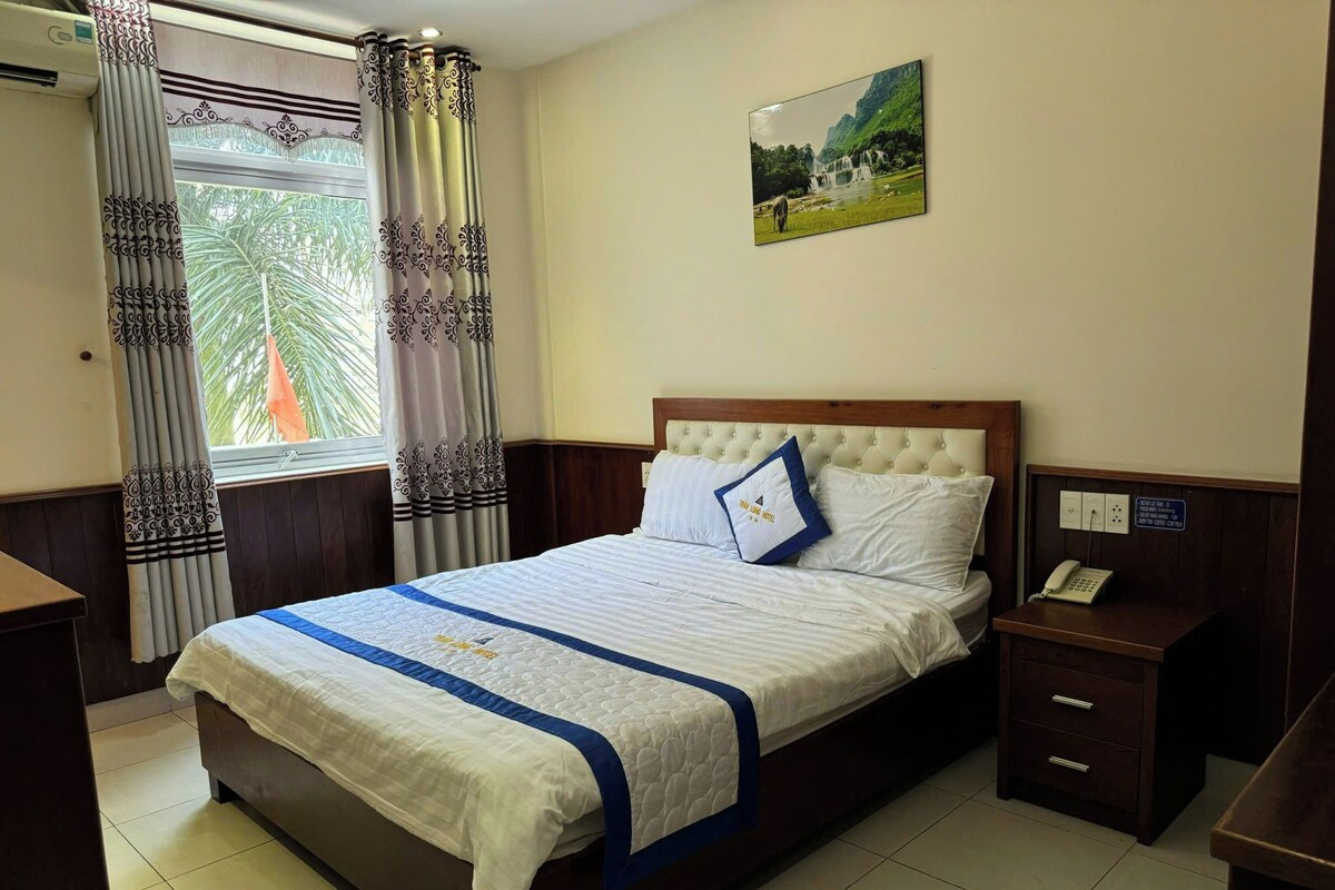 A hotel room with a bed, a nightstand, a telephone, a window with curtains, and a painting on the wall.
