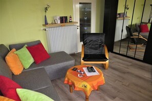A living room with a grey sofa, colorful cushions, a wooden chair, a small table with items, and a radiator.
