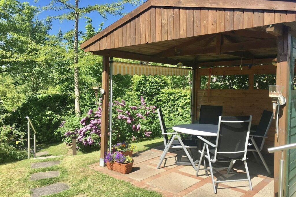 Un belvédère en bois, avec une table et des chaises, entouré d’un jardin agrémenté de plantes fleuries et d’un sentier en pierre.