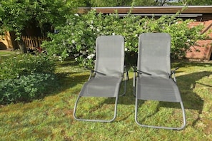 Two metal-framed chairs with mesh seats and backs, placed on grass in a garden with a wooden fence and flowering bushes in the background.