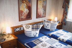 A bedroom with a wooden headboard, a bed with blue bedding and anchor-themed pillows, a nightstand with a lamp, and two paintings on the wall.
