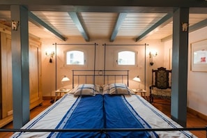 Una cama doble con colcha azul y blanca, dos lámparas de noche, una silla de madera y una mesita.