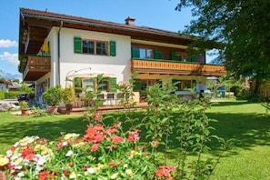 A two-story building with a balcony, a garden with flowers, and a clear sky. - Alpine view from the Thiele house (Schönau am Königssee)
