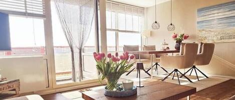 A modern living room with a wooden coffee table, a dining area with chairs, and a large window with a view.