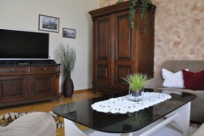 Ein Wohnzimmer mit einem Flachbildfernseher, einer Holzkommode, einem Glastisch mit einer Spitzendecke, einem Sofa und einem roten Kissen.