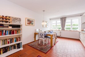 A room with a dining table, chairs, a bookshelf, a window with curtains, and a radiator. - Holiday apartment with 1 bedroom (Krummhörn-Greetsiel)