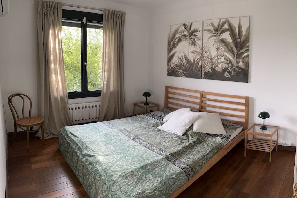 Ein Schlafzimmer mit einem Bett, einem Sessel, einem Fenster mit Vorhängen, einem Nachttisch, einer Lampe und einem Wandbild.