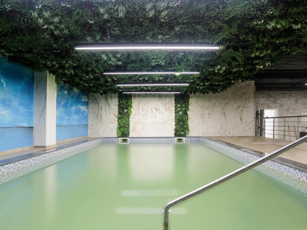 Una piscina interior con agua de color verdoso, rodeada de paredes con textura de hojas verdes y un techo con luminarias horizontales.