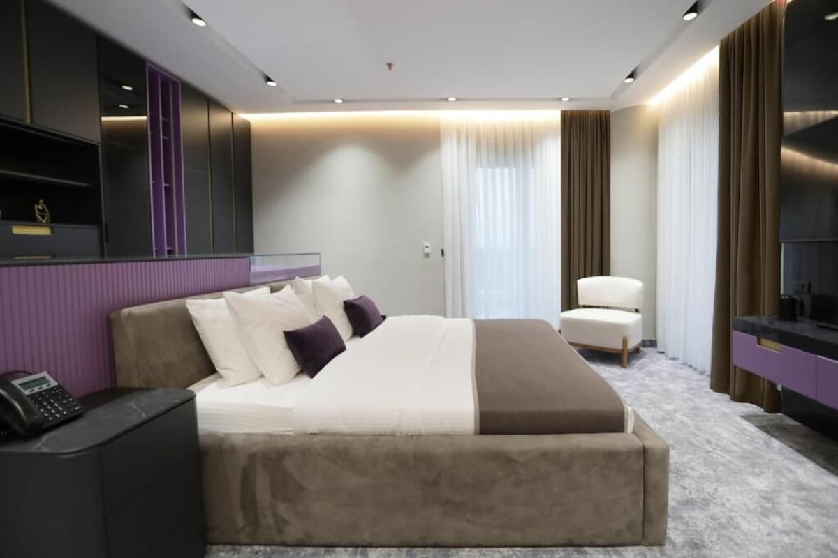 A modern hotel room with a large bed, a bedside table with a phone, a chair, and a TV mounted on the wall.