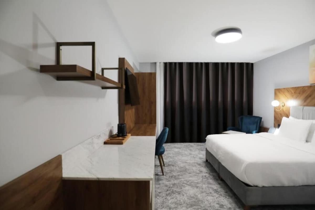 A hotel room with a bed, a desk, a chair, a coffee maker, and a large window with curtains.