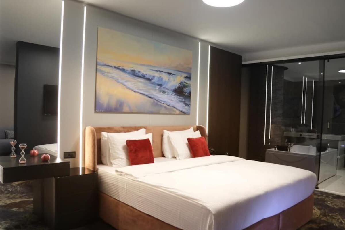 A hotel room with a large bed, a painting on the wall, a glass shower enclosure, and a bedside table with a lamp.