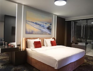 A hotel room with a large bed, a painting on the wall, a glass shower enclosure, and a bedside table with a lamp. - Hotel Gallery  (Gjilan)