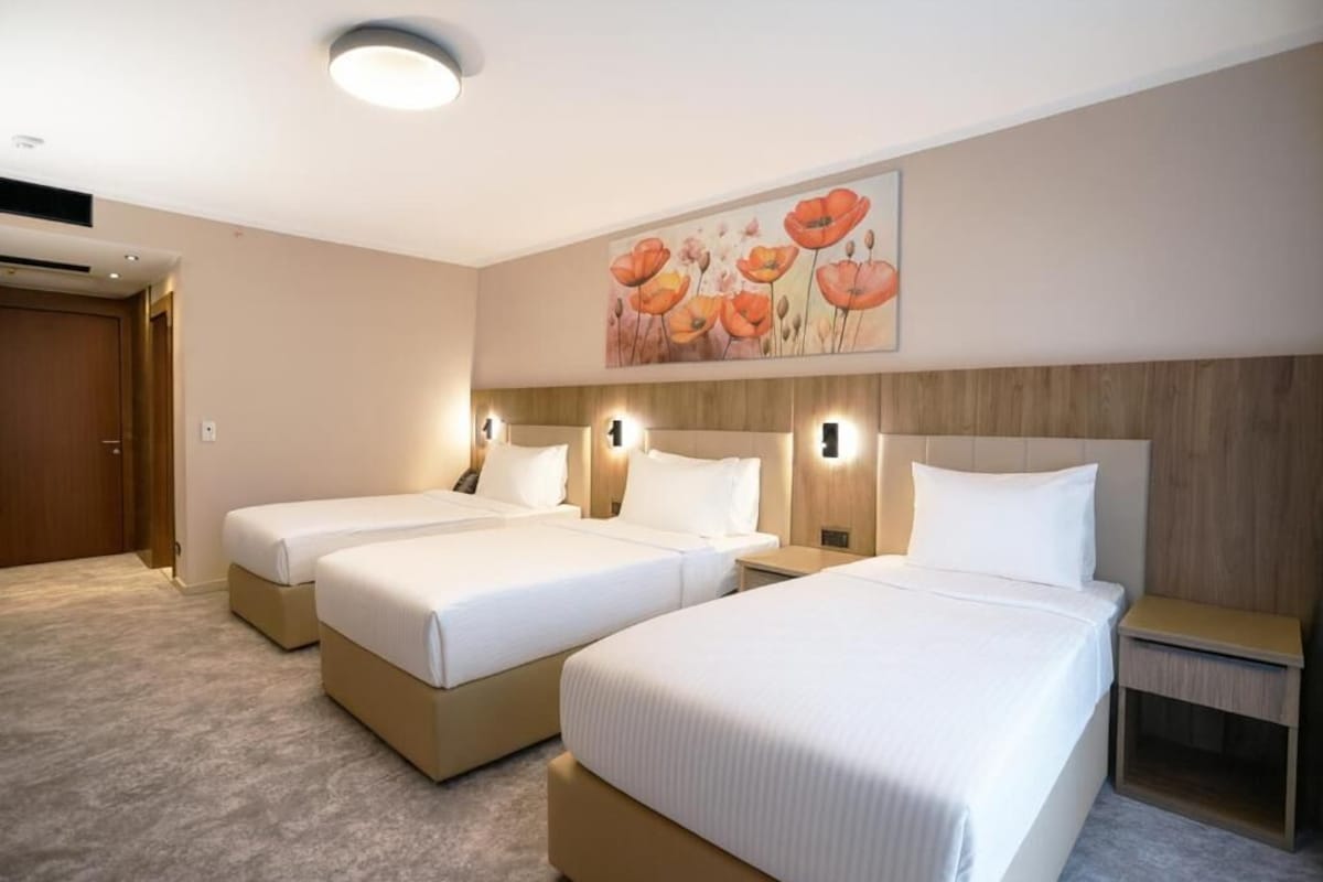 A hotel room with three beds, a wooden headboard, a painting of flowers, and a carpeted floor.