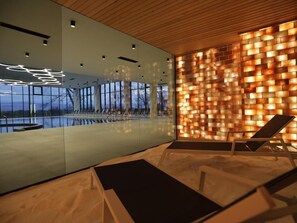 Un área de piscina interior moderna con sillones de descanso, un muro de cristal que refleja la piscina, y una pared de ladrillo con iluminación.