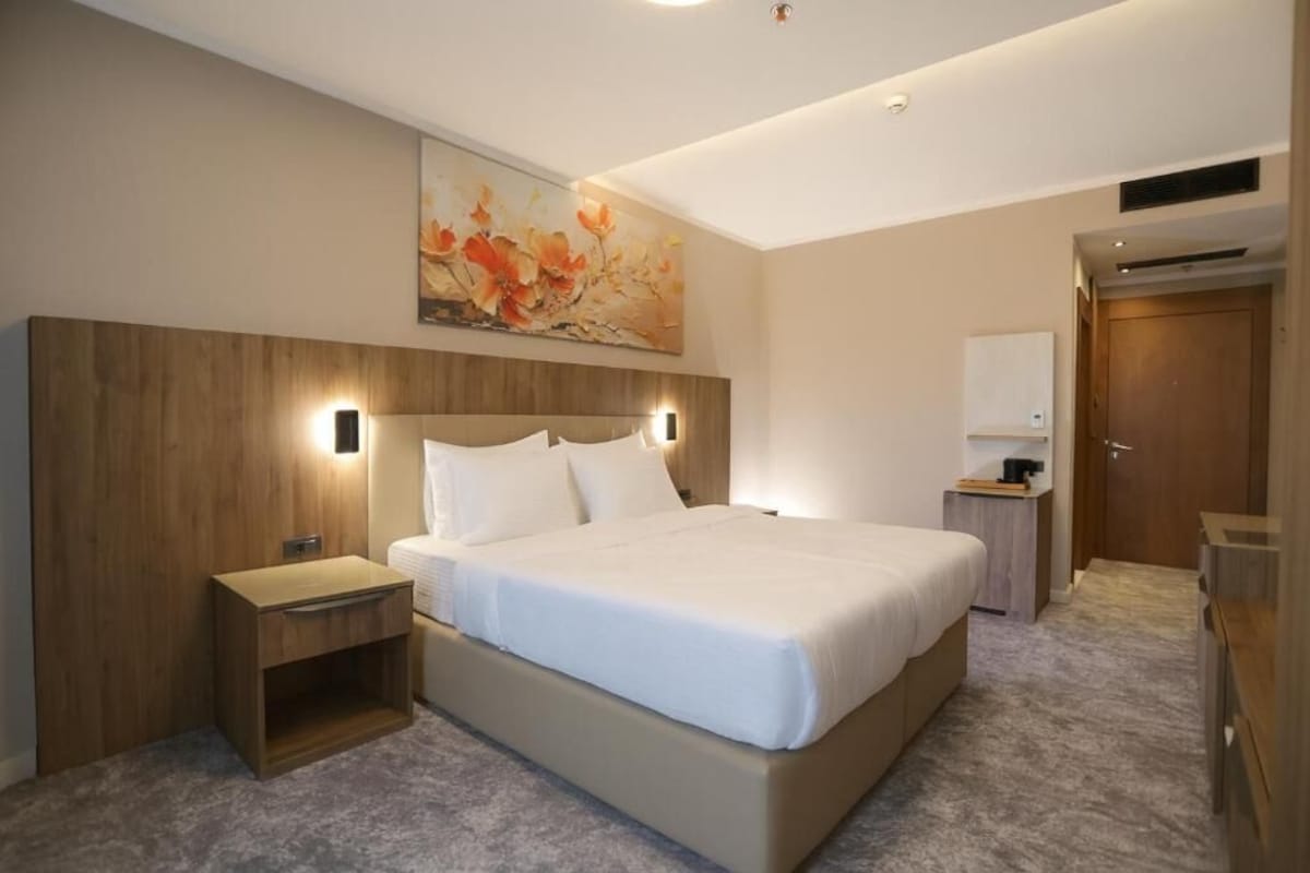 A modern hotel room with a large bed, a wooden headboard, a nightstand, a bedside table, and a painting on the wall.