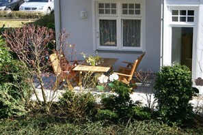 Ein Wohnhaus mit einer Terrasse, die einen Holztisch und Stühle sowie verschiedene Sträucher und einen kleinen Baum umfasst.