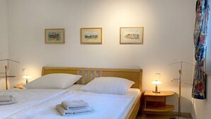 Un llit doble amb roba de llit blanca i dos almoiners, una taula de nit amb una làmpada, i tres quadres emmarcats a la paret.
