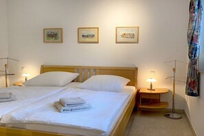 Een tweepersoonsbed met witte lakens en twee kussens, een nachtkastje met lamp, en drie ingelijste kunstwerken aan de muur.