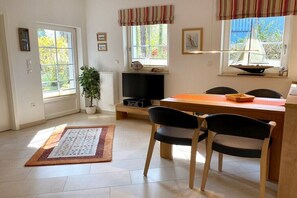 Een kamer met een eettafel, stoelen, een televisie, een kamerplant en een raam met een model zeilboot.