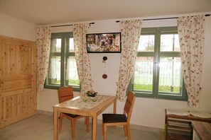 Ein Zimmer mit Holztisch und -stühlen, einem an der Wand befestigten Fernseher und großen Fenstern mit blumigen Vorhängen.