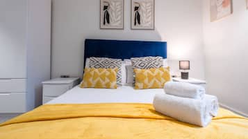 Una cama bien hecha con una manta amarilla, dos almohadas con diseños geométricos y una toalla blanca doblada al lado.