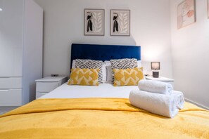 Una cama bien hecha con una manta amarilla, dos almohadas con diseños geométricos y una toalla blanca doblada al lado.