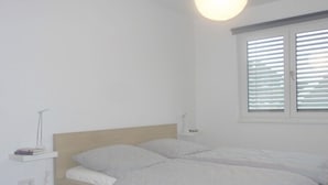 A single bed with a wooden headboard, a bedside table with a lamp, and a window with blinds. - Holiday apartment Husumer Wattwurm (Husum)