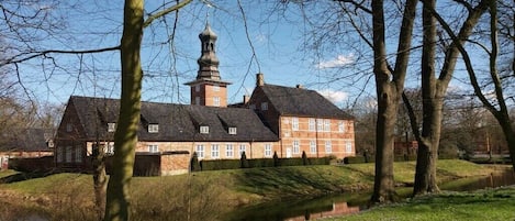 A large, historic building with a prominent tower, surrounded by trees and a small body of water.