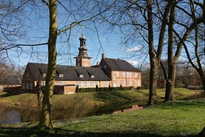 Un grande edificio storico con una torre imponente, circondato da alberi e un piccolo specchio d'acqua.