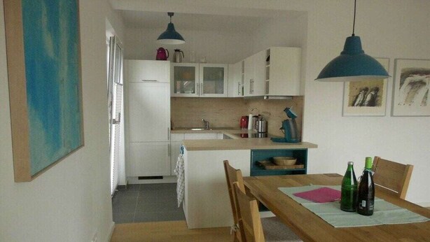 A compact kitchen with white cabinets, a wooden dining table, and two pendant lights. - Holiday apartment Husumer Wattwurm (Husum)