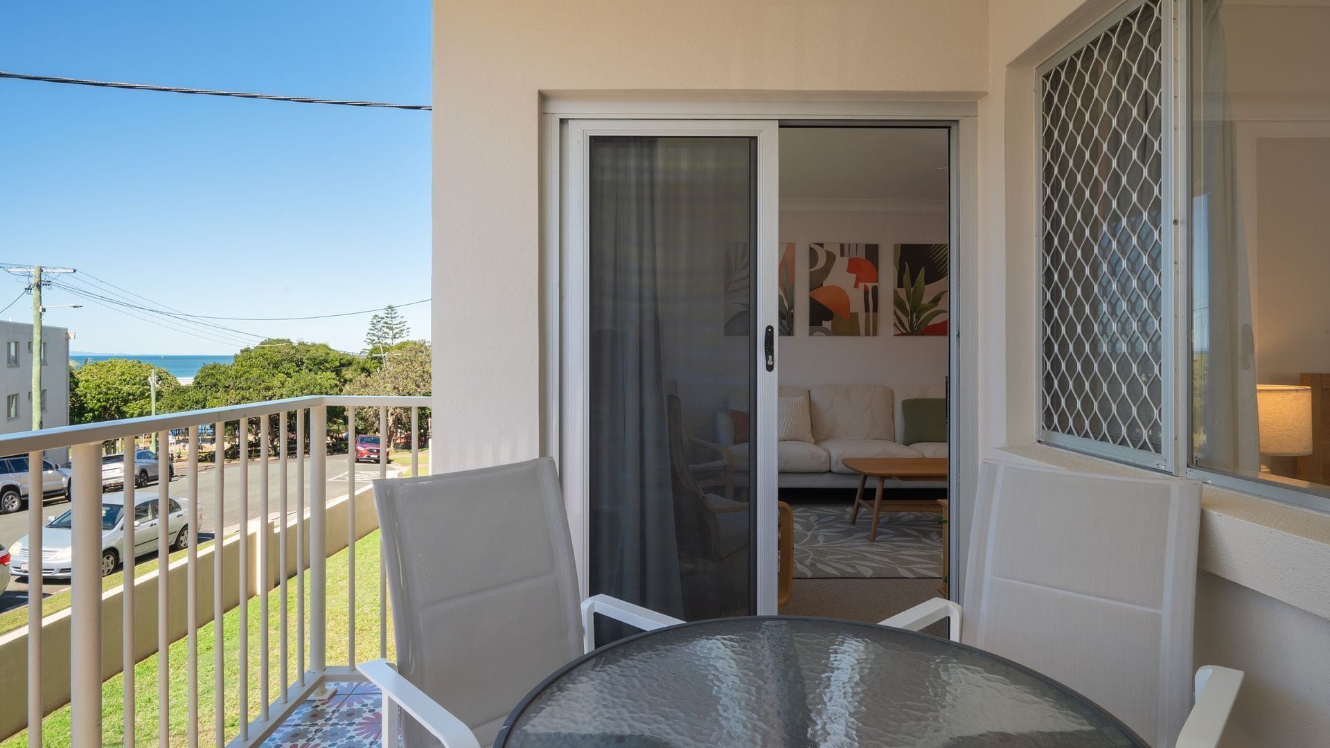 A balcony with a glass table and chairs, a sliding glass door leading to a living area with a sofa and a coffee table, and a view of the street and ocean.