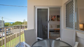 A balcony with a glass table and chairs, a sliding glass door leading to a living area with a sofa and a coffee table, and a view of the street and ocean. - Star Lodge unit 6 Bulcock Beach QLD (Caloundra)