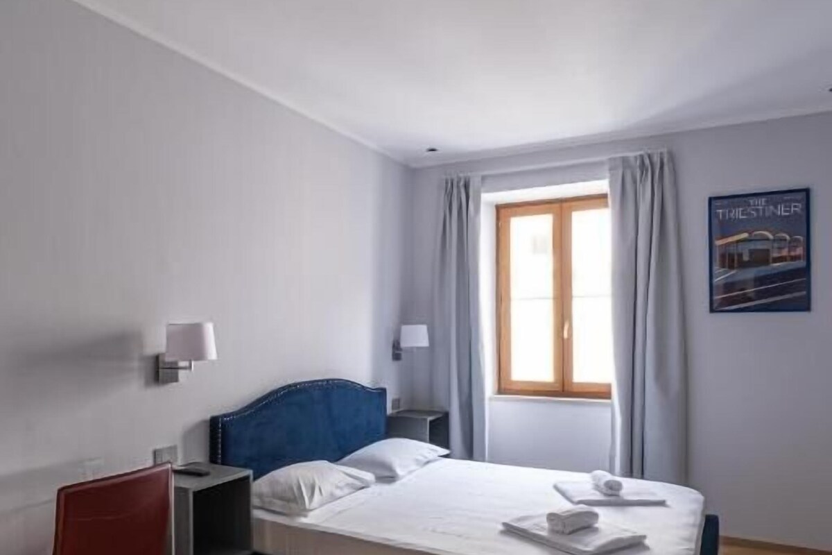 Ein Schlafzimmer mit Bett, Schreibtisch, Stuhl, Nachttisch, einem Fenster mit Vorhängen und einer Wandlampe.