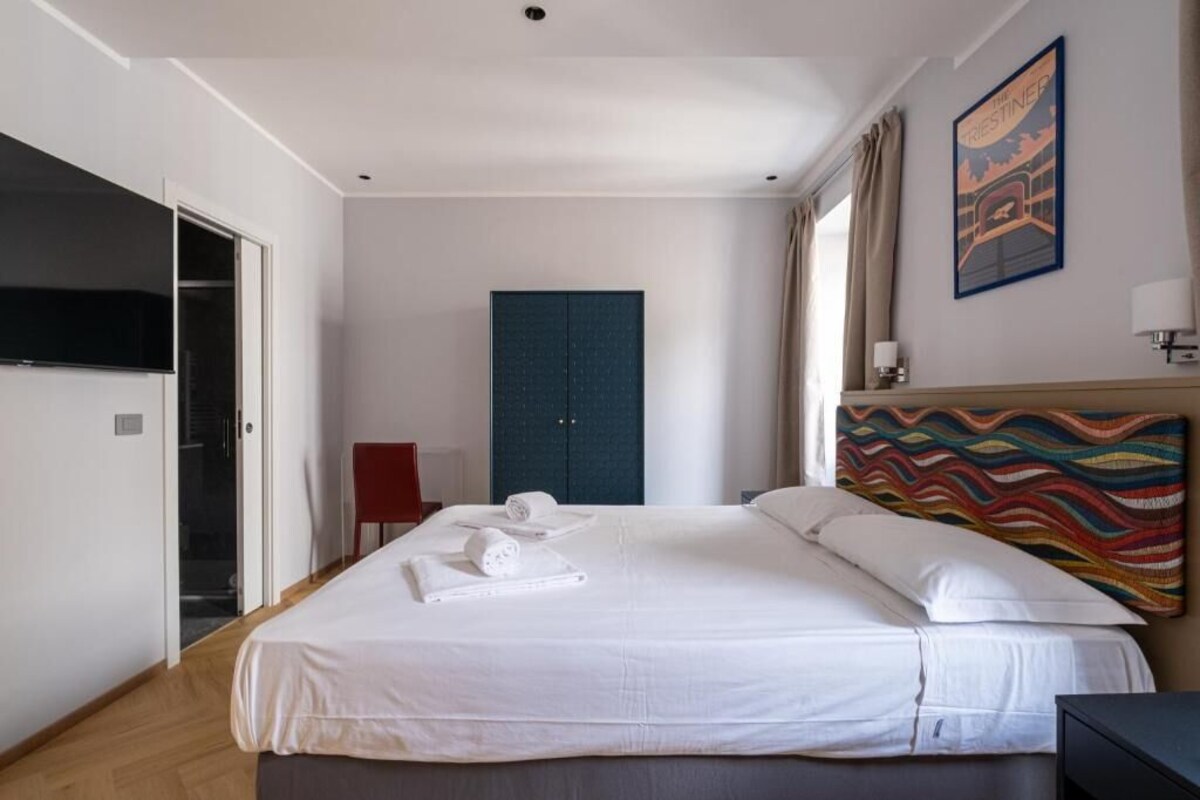 Ein Hotelzimmer mit einem Bett, einem Kleiderschrank, einem roten Sessel, einem Fernseher und einem farbenfrohen Kunstwerk an der Wand.