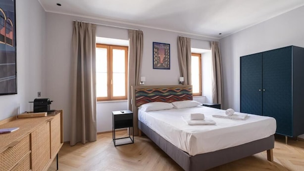 A hotel room with a bed, a wardrobe, a desk, and a window with curtains. - 23 SUITES (TRIESTE)