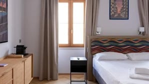 A hotel room with a bed, a nightstand, a window with curtains, a wall-mounted artwork, and a wooden floor. - 23 SUITES (TRIESTE)