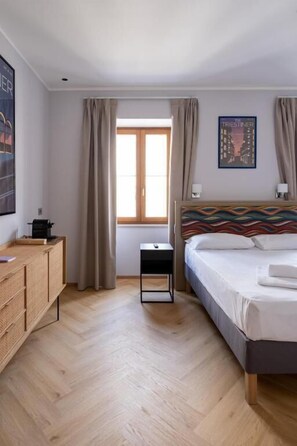 Ein Hotelzimmer mit Bett, Nachttisch, einem Fenster mit Vorhängen, einem Wandbild und Holzboden.