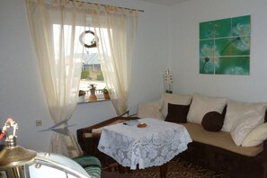 A room with a sofa, a table with a lace tablecloth, a window with sheer curtains, and a wall-mounted artwork. - Holiday home with 2 bedrooms (Waren\/Müritz)