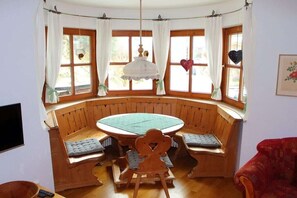 Ein Zimmer mit einem runden Holztisch, Bänken, großen Fenstern mit Vorhängen, einer Hängelampe und einem roten Stuhl.