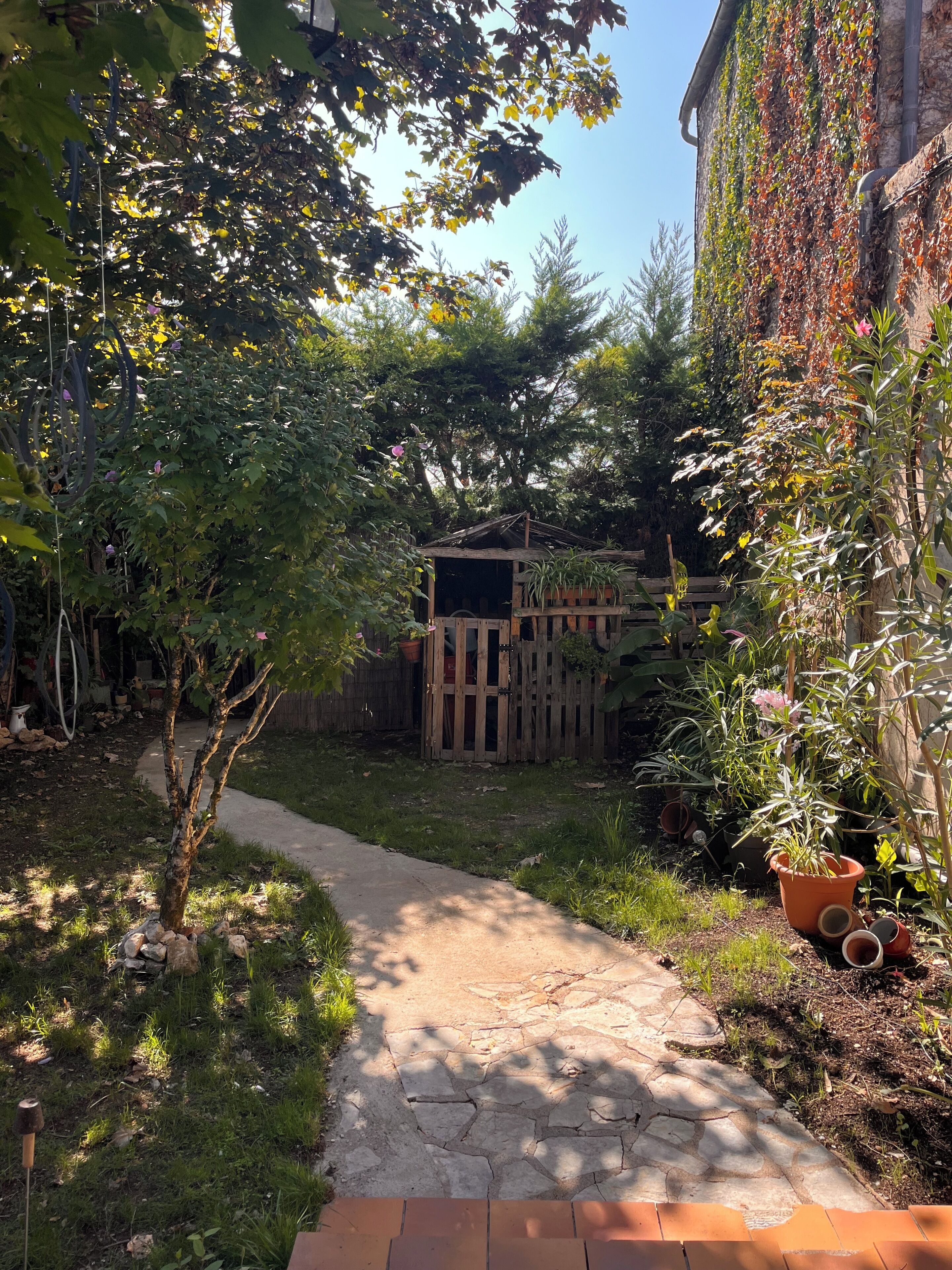 Ein Gartenpfad führt zu einer Holzbeschreibung, umgeben von Pflanzen und einer Ziegelwand.